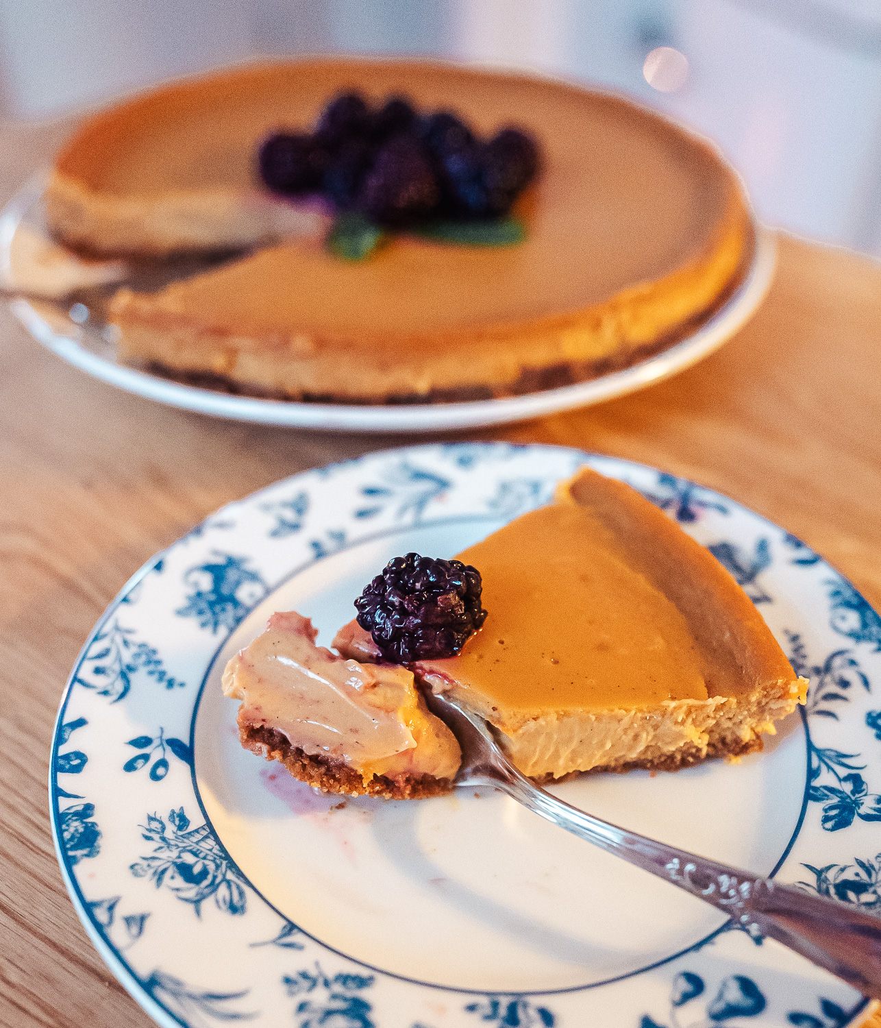 slice of Norwegian brunost cheesecake with a blackberry on top served on a white plate with a blue design on the edges